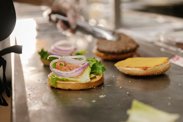 Two open burger buns with onions and lettuce sit on the counter with employee putting patties on them